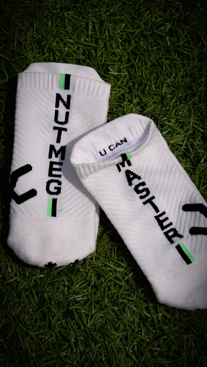 A pair of white low-cut athletic socks with black and green graphic details, laid flat on a dark green grass background. One sock features the vertical text 'NUTMEG' and the other features 'U CAN' alongside a black graphic.