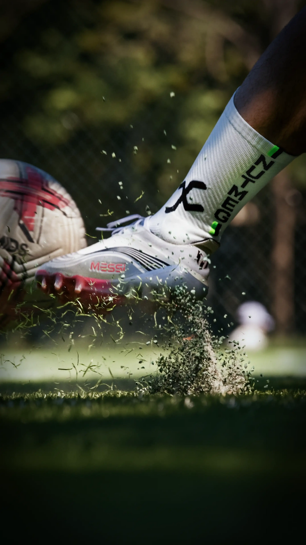 A close-up action shot of a soccer player's foot striking a ball on a grass field, wearing a white soccer cleat with red accents, a white sock with black and green lettering, and a dark athletic jersey.