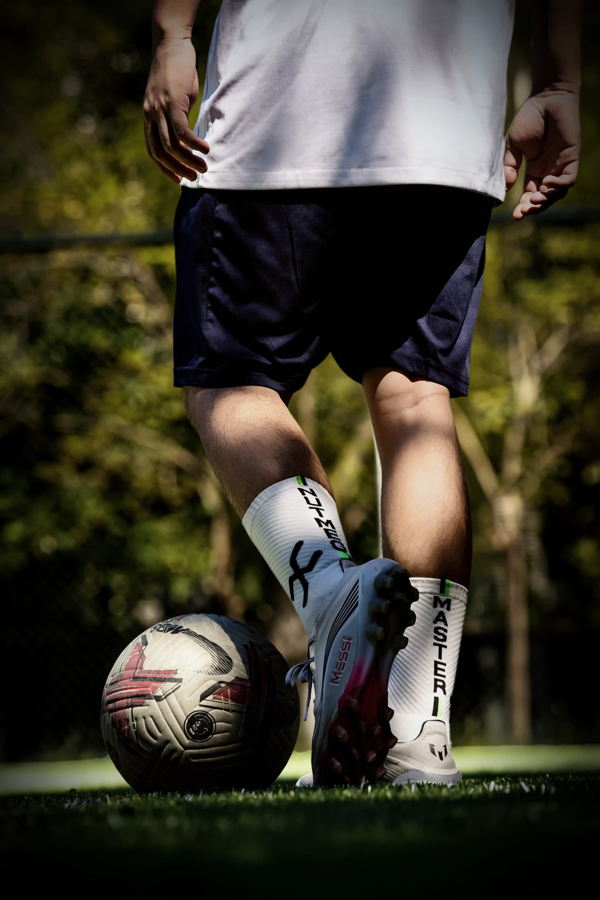 A person outdoors on grass wearing white soccer socks with black text, white soccer cleats, and navy shorts, with a soccer ball on the grass nearby.