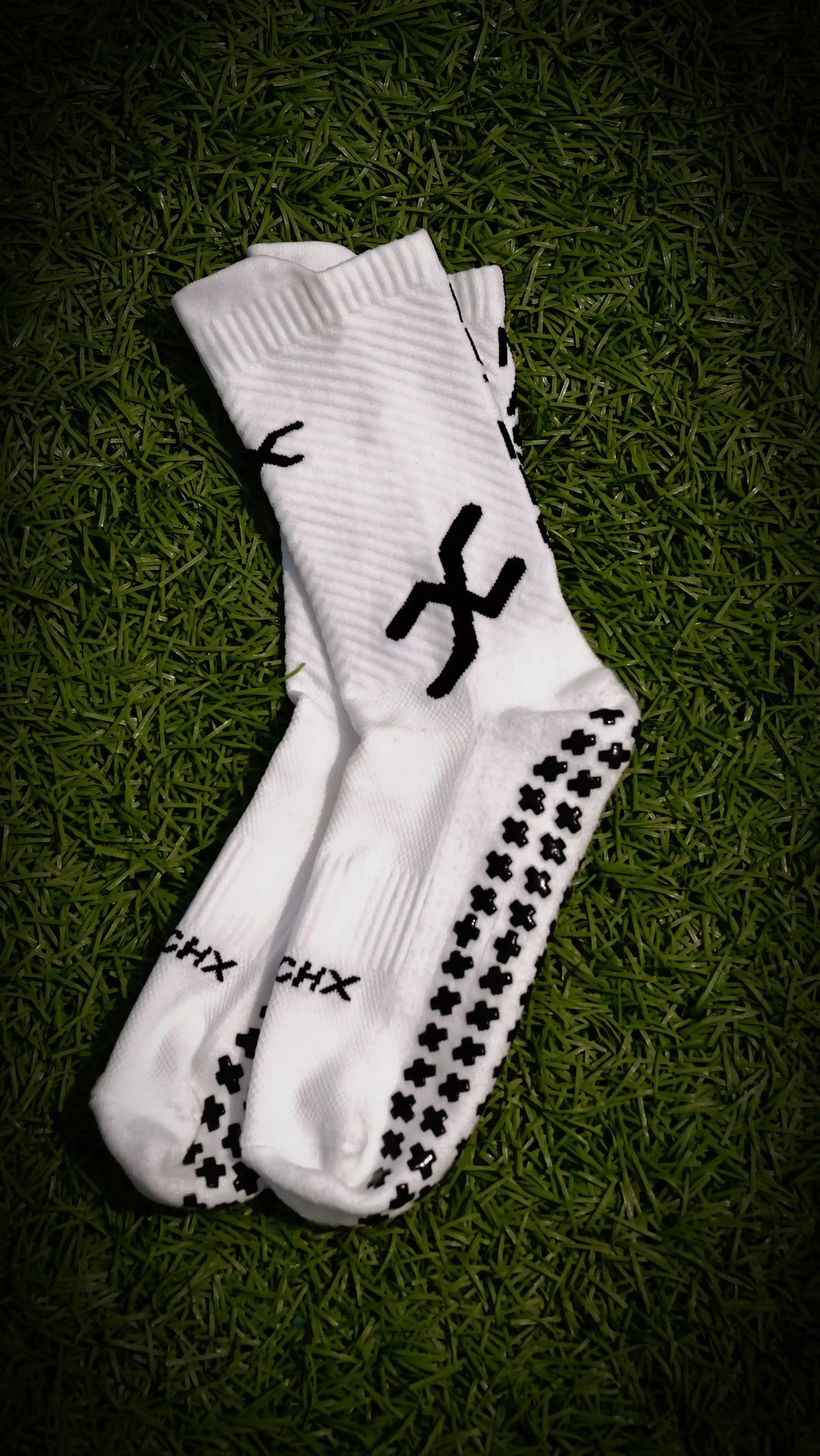 A pair of white athletic crew socks with black grip dots and logo graphics, laid flat on a dark green grass background.