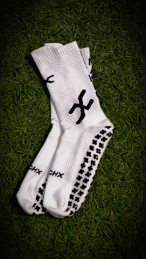 A pair of white athletic crew socks with black grip dots and logo graphics, laid flat on a dark green grass background.
