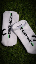 A pair of white low-cut athletic socks with black and green graphic details, laid flat on a dark green grass background. One sock features the vertical text 'NUTMEG' and the other features 'U CAN' alongside a black graphic.