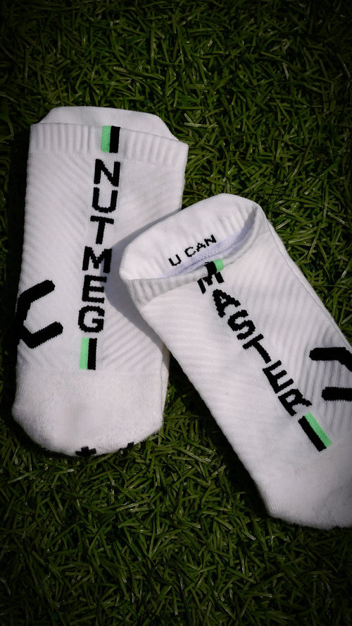 A pair of white low-cut athletic socks with black and green graphic details, laid flat on a dark green grass background. One sock features the vertical text 'NUTMEG' and the other features 'U CAN' alongside a black graphic.